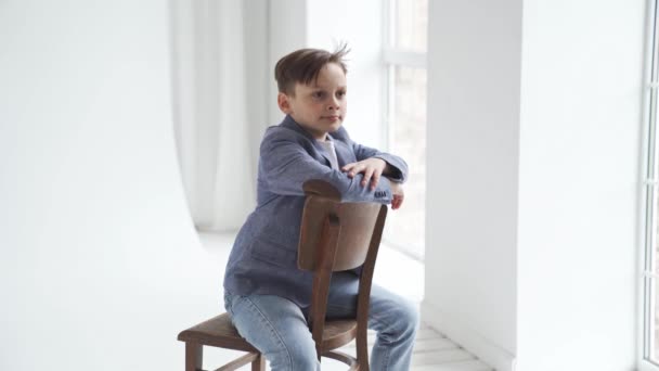 Un adolescent dans une veste et un jean pose sur une chaise par une fenêtre. — Video