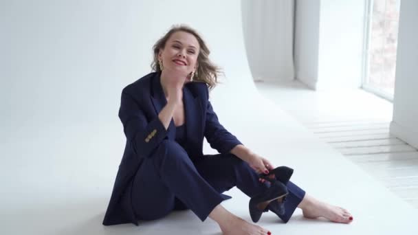 Woman in blue suit sits barefoot on in white photo studio and holds heeled shoes — Stock Video