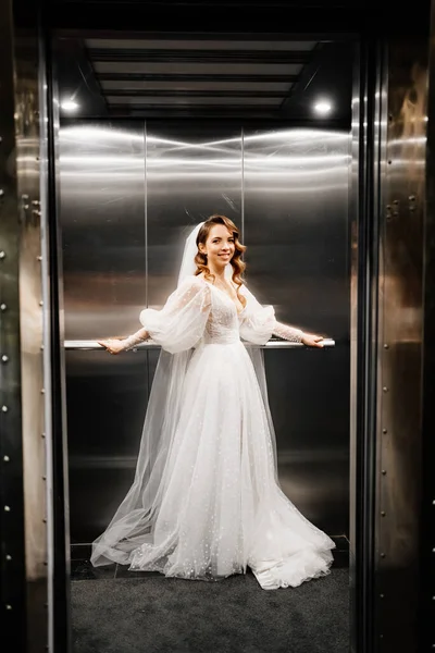 Bruid in een witte elegante jurk, met een lange sluier staat in een donkere lift. — Stockfoto