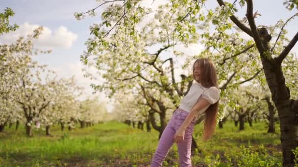 Vrolijk tiener meisje met lang haar wandelingen in een open plek tussen bloeiende bomen — Stockvideo