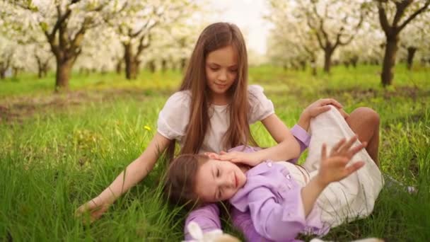 Två långhåriga flickor systrar på gräsmattan i trädgården med blommande träd. — Stockvideo