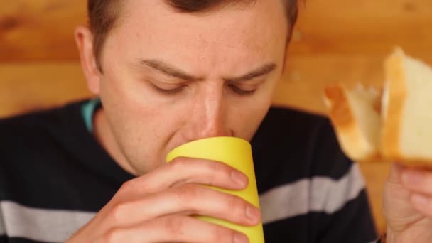 Un uomo mangia un panino fatto di pane bianco e bevande da un bicchiere. — Video Stock