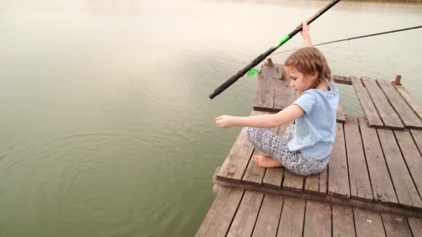 Niña se sienta con caña de pescar capturado pequeño pez en puente por río. — Vídeos de Stock