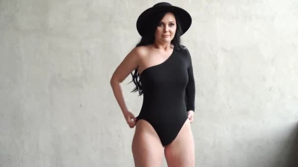 Piękna i seksowna kobieta w czarnym body i kapeluszu na szarej ścianie. — Wideo stockowe