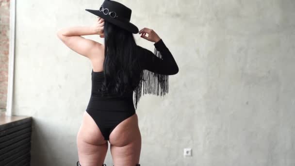 Vista posteriore. Bella e sexy donna in un body nero e cappello al muro grigio — Video Stock