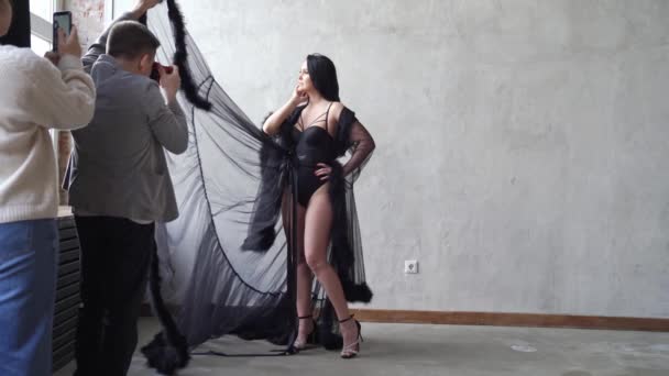 Mulher sexy em bodysuit e negligee fica na parede cinza. bastidores sessão de fotos — Vídeo de Stock