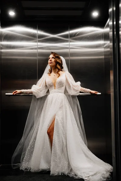 Невеста в белом элегантном платье, с длинной завесой стоит в темном лифте. — стоковое фото