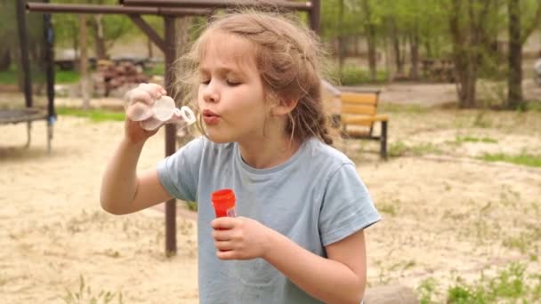 Egy kis bozontos lány kék pólóban felfújja a szappanbuborékokat a parkban. — Stock videók