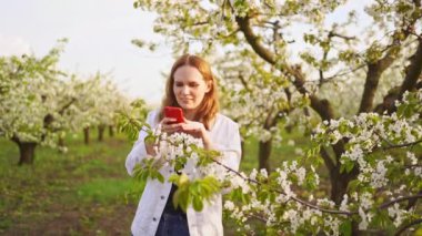 Beyaz ceketli bir kadın çiçeklerle dolu bir bahçede telefonuyla video çekiyor..