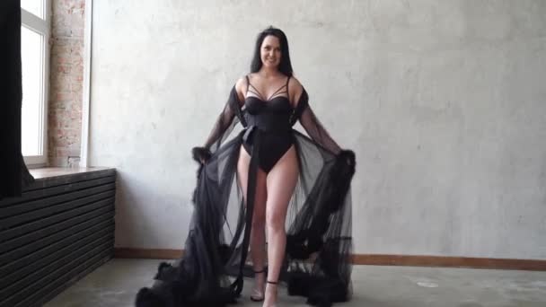 Mujer sexy en traje de cuerpo negro y negligee se encuentra en la pared gris. sesión de fotos — Vídeo de stock