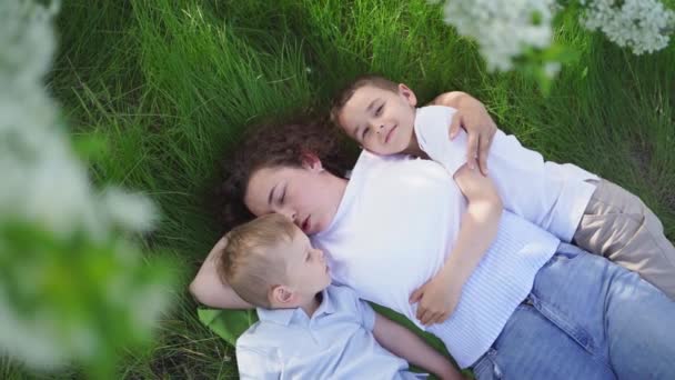 Eine Mutter mit zwei kleinen Söhnen liegt unter den Ästen eines blühenden Baumes. Picknick — Stockvideo