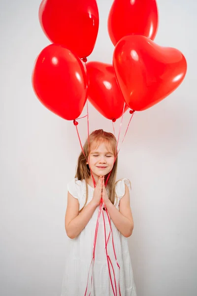 Fille drôle avec des ballons rouges souriant, plie les paumes en face de près de lui mur blanc — Photo