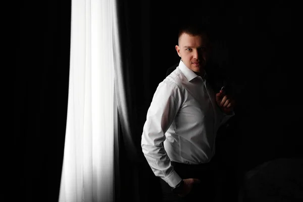 Mann in weißem Hemd mit Jacke in der Hand am Fenster in einem dunklen Raum. — Stockfoto