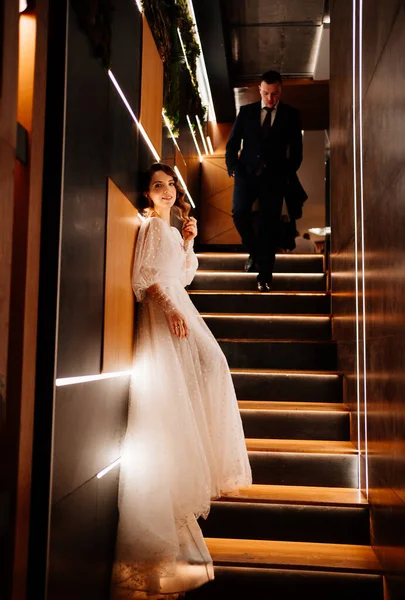 Der Bräutigam im Anzug und die Braut im weißen Kleid stehen auf der schlecht beleuchteten Treppe — Stockfoto