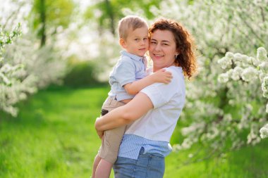 Anne küçük oğlunu çiçek açan bir bahar bahçesinde tutuyor. Ebeveyn sevgisi ve bakımı