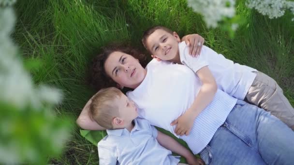 Una madre con dos hijos pequeños yace bajo las ramas de un árbol floreciente. picnic — Vídeo de stock