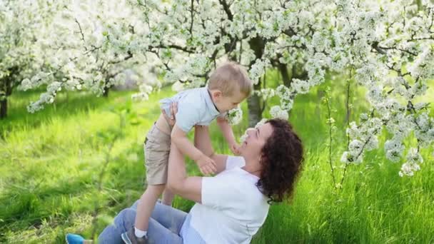 Mama bawi się w wiosennym ogrodzie z młodym synem, rzuca dziecko w górę. — Wideo stockowe