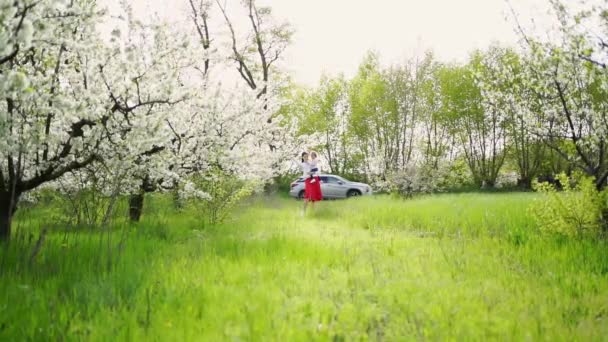 Ibu dengan anak perempuan dalam pelukannya di taman dengan pohon berbunga pergi ke mobil. — Stok Video