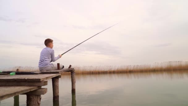 Un ragazzo siede su un ponte di legno e cattura un pesce sulla canna da pesca. — Video Stock
