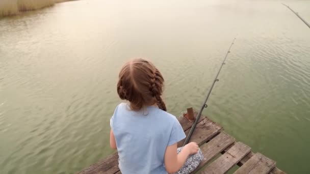 Маленька дівчинка сидить з вудкою на мосту біля річки . — стокове відео