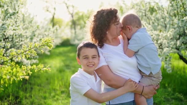 Мать с сыновьями гуляет в цветущем саду. Младший ребенок на руках, зевает — стоковое видео