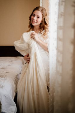 Gelin beyaz ipek bornozlu, pencerede gelinlik var.