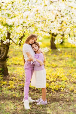 İki kız kardeş bahçede çiçek açan ağaçlarla eğlenip kucaklaşıyorlar..