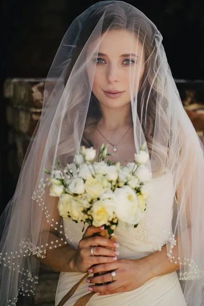 Hermosa novia con el pelo ondulado largo bajo el velo exterior con un ramo de flores —  Fotos de Stock