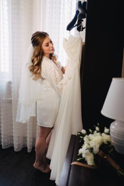 Gelin beyaz ipek bornozla gelinliğin yanında askıda asılı duruyor.. 