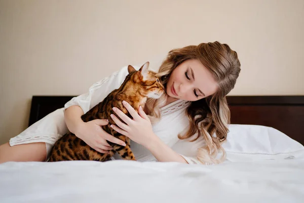 Schöne junge Frau in einem weißen Seidenmantel auf einem Bett mit einer gestreiften Katze. — Stockfoto