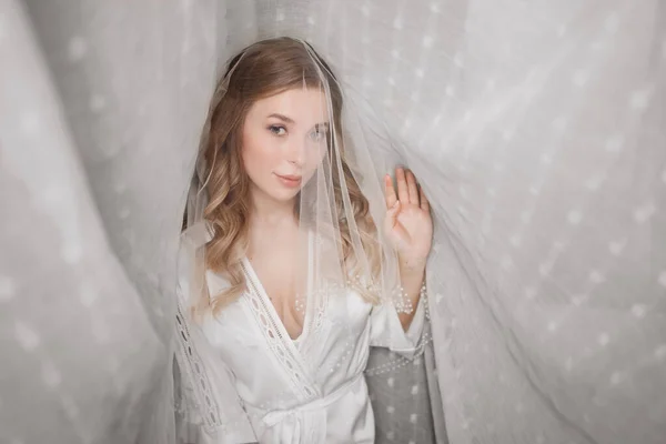 En mild morgon för bruden. vacker ung kvinna i slöja under en vit gardin — Stockfoto