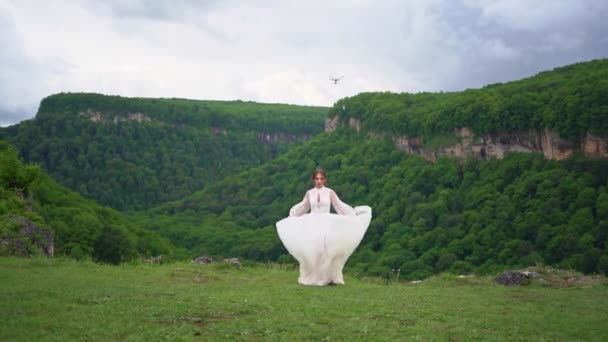 Egy lány menyasszonyi ruhában és fekete csizmában táncol a füvön a hegyekben.. — Stock videók