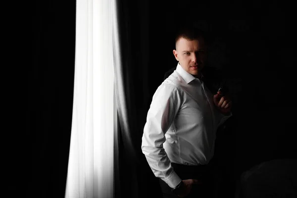 一个穿着白衬衫，手里拿着一件夹克的男人在一间黑暗的房间的窗边. — 图库照片