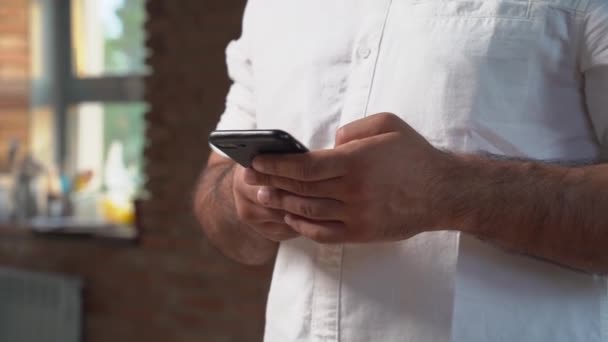 Een man in een wit shirt houdt een smartphone vast en schrijft een boodschap. — Stockvideo