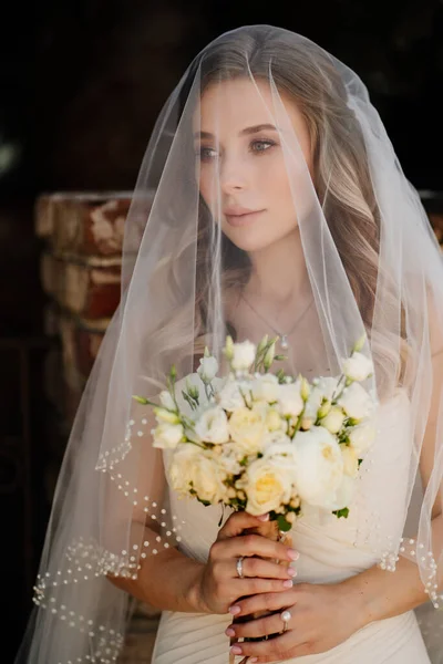 Hermosa novia con el pelo ondulado largo bajo el velo exterior con un ramo de flores —  Fotos de Stock