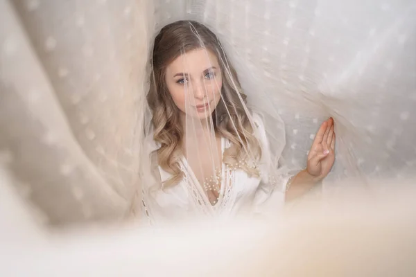 La mañana tierna de la novia. hermosa joven en velo bajo una cortina blanca — Foto de Stock