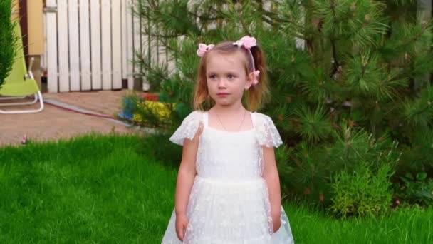 Красивая маленькая девочка в белом платье стоит и говорит что-то на зеленом газоне — стоковое видео