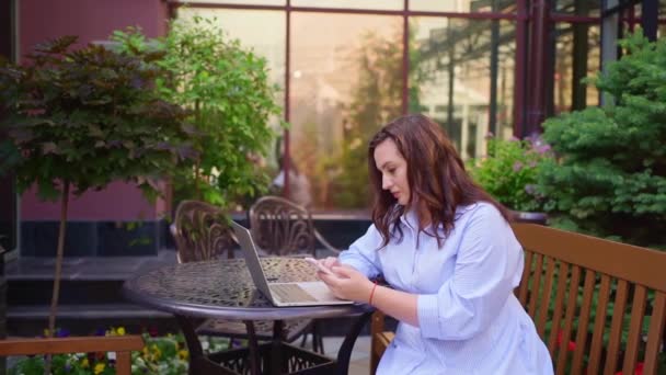 La donna si siede su una terrazza all'aperto e lavora su un computer portatile e uno smartphone. — Video Stock