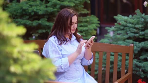 Die Frau sitzt auf einer Bank im Garten und schreibt SMS auf ihrem Smartphone. — Stockvideo
