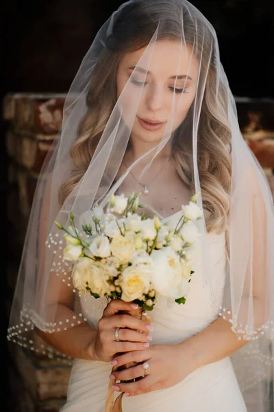 ベールの下に長い波状の髪をした美しい花嫁を花束で外に — ストック写真