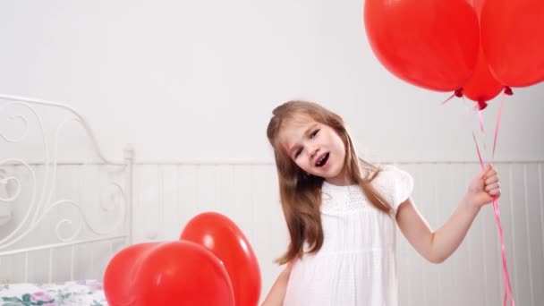 Rolig flicka med röda ballonger i form av hjärtan ler på sängen av vit vägg — Stockvideo