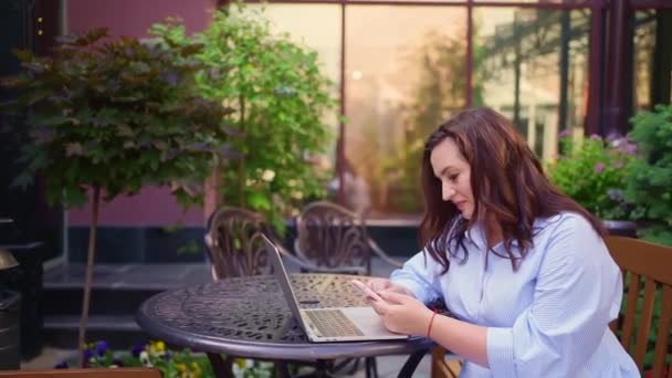 Kvinnan sitter på en utomhusterrass och arbetar på en laptop och en smartphone. — Stockvideo