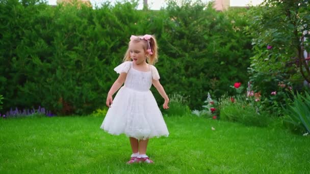 Una hermosa niña en un vestido blanco se encuentra en el césped verde en el jardín. — Vídeos de Stock