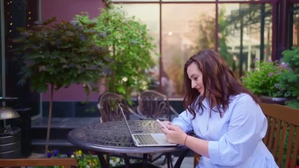 La donna si siede su una terrazza all'aperto e lavora su un computer portatile e uno smartphone. — Video Stock