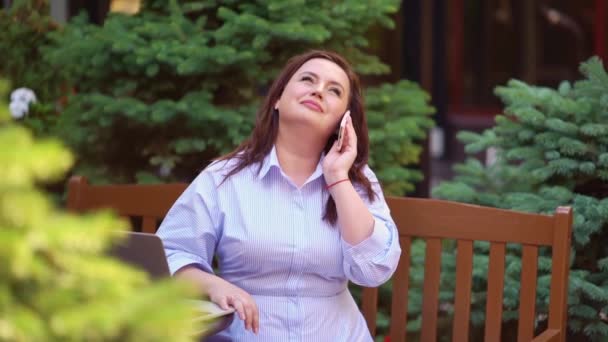 A mulher se senta em um banco no jardim e fala em seu smartphone. — Vídeo de Stock