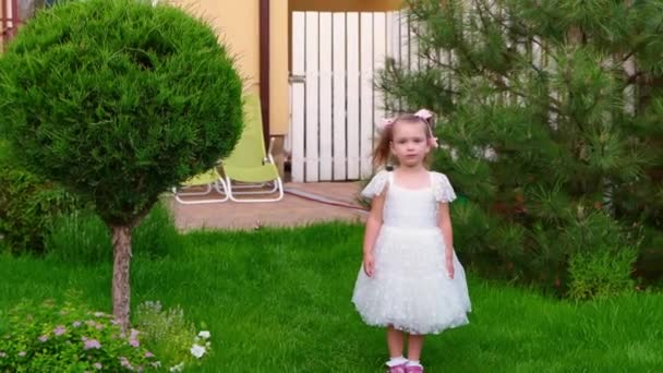 Ein schönes kleines Mädchen im weißen Kleid steht auf dem Rasen und schüttelt den Kopf. — Stockvideo