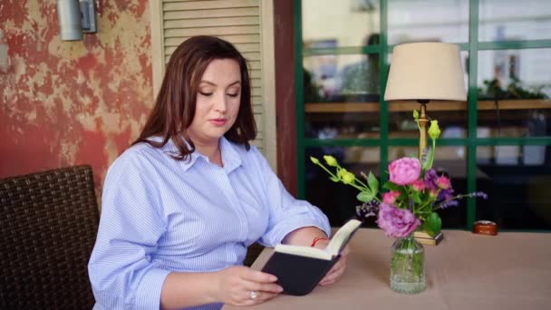 Una mujer se sienta en una mesa en un café o en la terraza y lee un libro en voz alta. — Vídeos de Stock