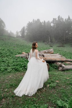 Beyaz gelinlik giymiş gelin kütüklerin yanındaki tarlada duruyor..
