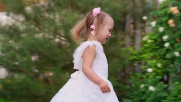 Feliz niña en un vestido blanco inteligente corre por el césped en el jardín. — Vídeo de stock