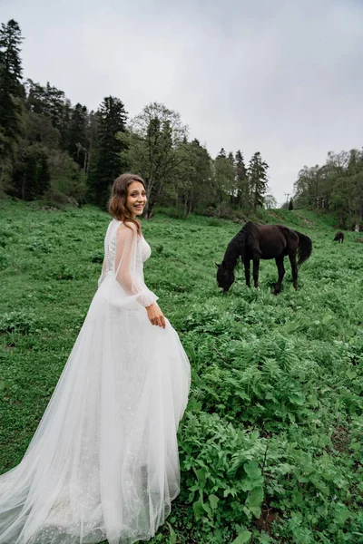 Uma jovem mulher bonita em um vestido de noiva branco fica em um campo com cavalos. — Fotografia de Stock